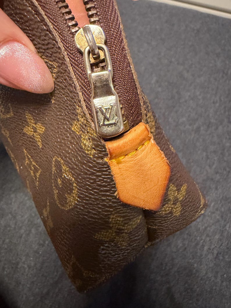 May include: Close-up of a brown and tan monogram canvas pouch with a gold-tone zipper and leather pull tab. The zipper pull features a silver-colored metal with the brand's logo. The pouch is a small, rectangular shape.