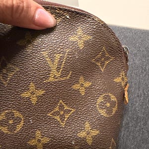 May include: Brown and tan cosmetic bag with a repeating pattern of interlocking logos. The bag has a curved top and a zipper closure. The bag is held by a hand.