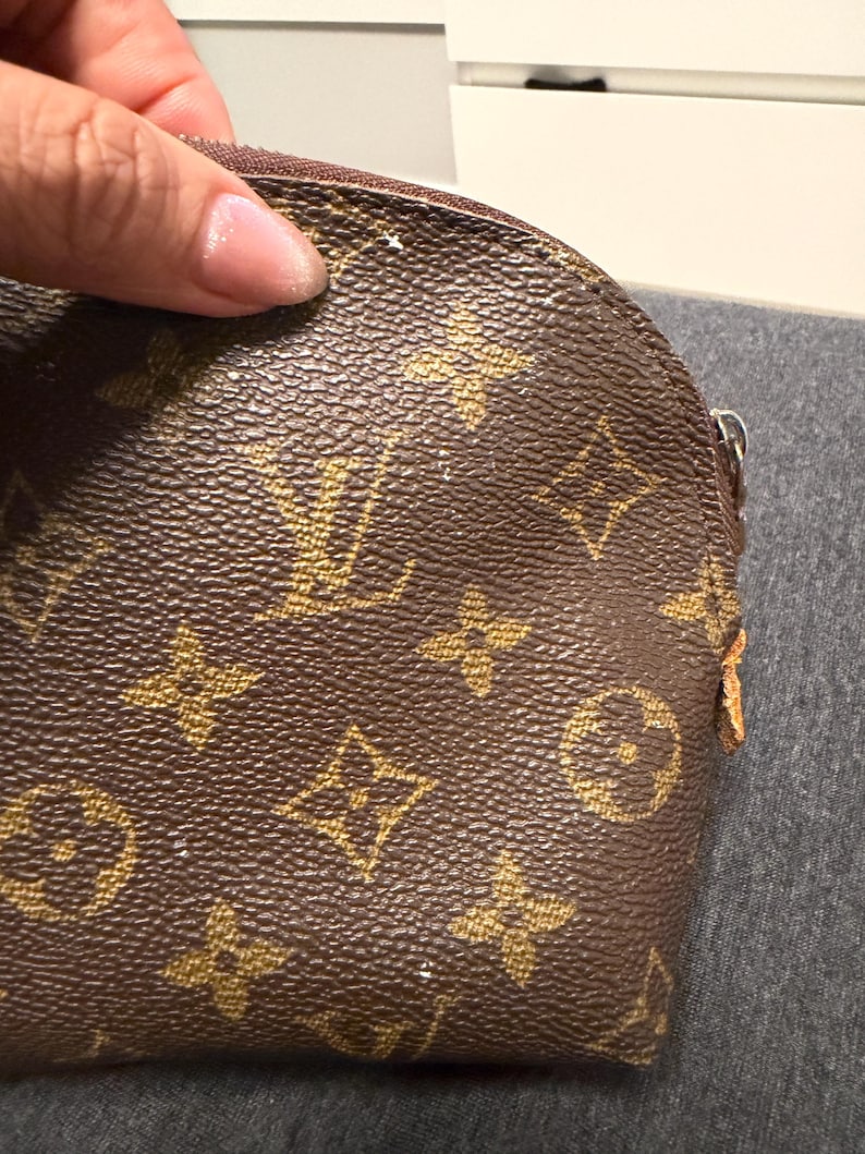 May include: Brown and tan cosmetic bag with a repeating pattern of interlocking logos. The bag has a curved top and a zipper closure. The bag is held by a hand.