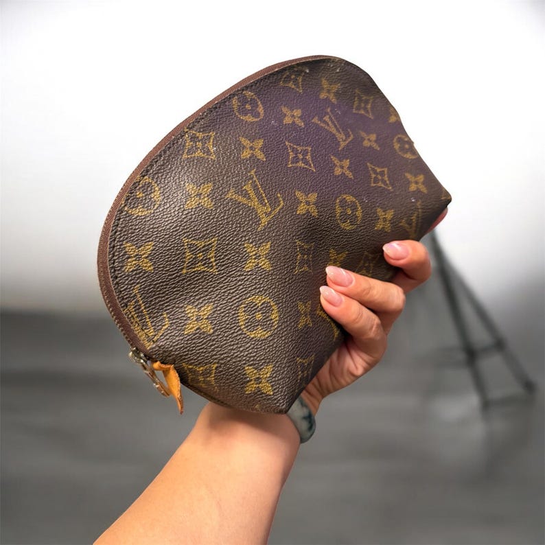 May include: Brown cosmetic bag with a repeating gold logo pattern. The bag has a curved shape, a zipper closure, and a leather pull tab. The bag is held in a person's hand.
