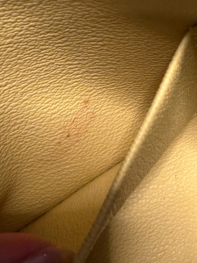 May include: Close-up of a light yellow leather interior with a textured surface. The leather has a subtle grain pattern and a small, reddish-brown stain. The image shows the inside of a wallet or similar item.