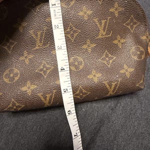 May include: Brown cosmetic bag with a repeating pattern of gold-colored logos. The bag is approximately 8 inches long. A white measuring tape is placed next to the bag.