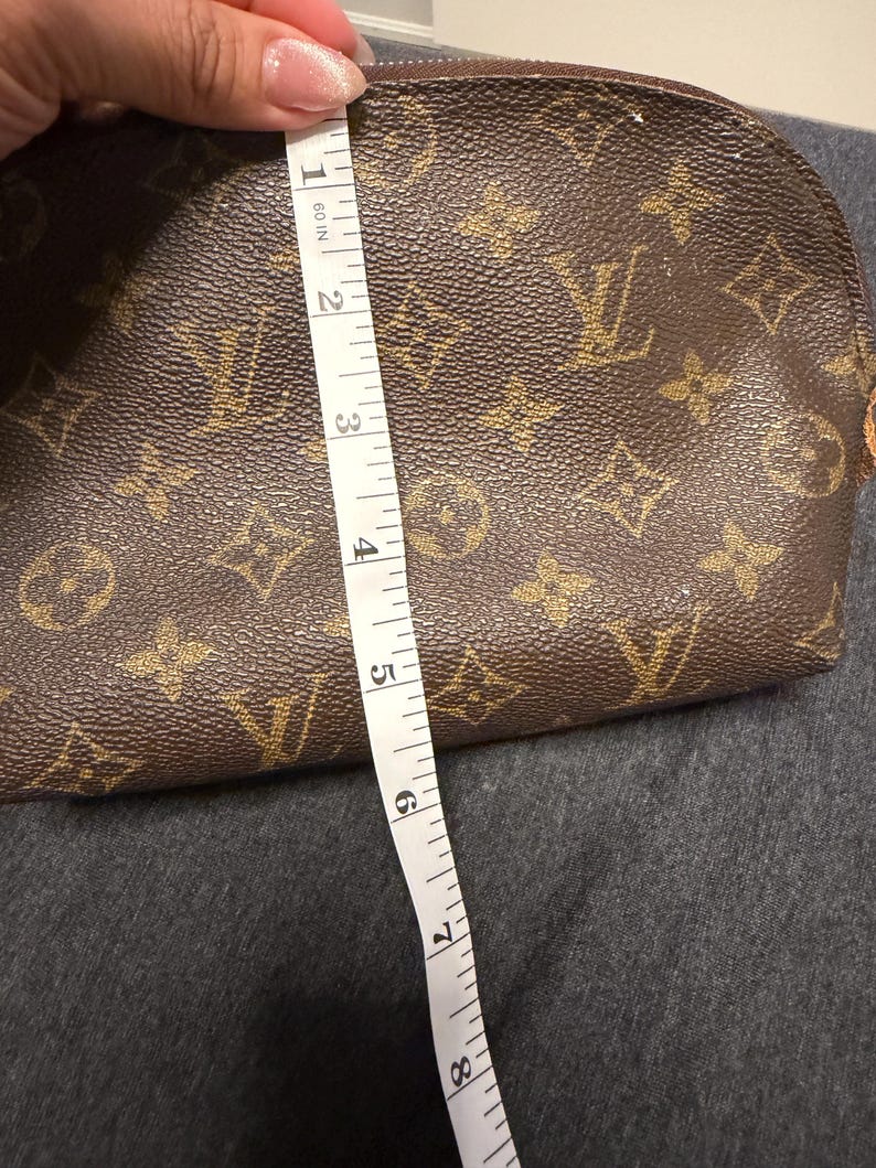 May include: Brown cosmetic bag with a repeating pattern of gold-colored logos. The bag is approximately 8 inches long. A white measuring tape is placed next to the bag.
