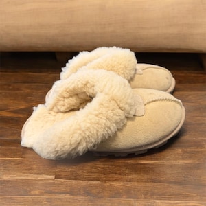 Peut inclure: Une paire de pantoufles en daim beige avec une doublure en peau de mouton crème. Les pantoufles ont un bout arrondi et une semelle en caoutchouc. La peau de mouton entoure le dessus du pied.