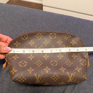 May include: Brown cosmetic bag with a repeating pattern of interlocking floral and logo designs. The bag is approximately 9 inches long. The zipper is brown, and the bag is on a gray surface.