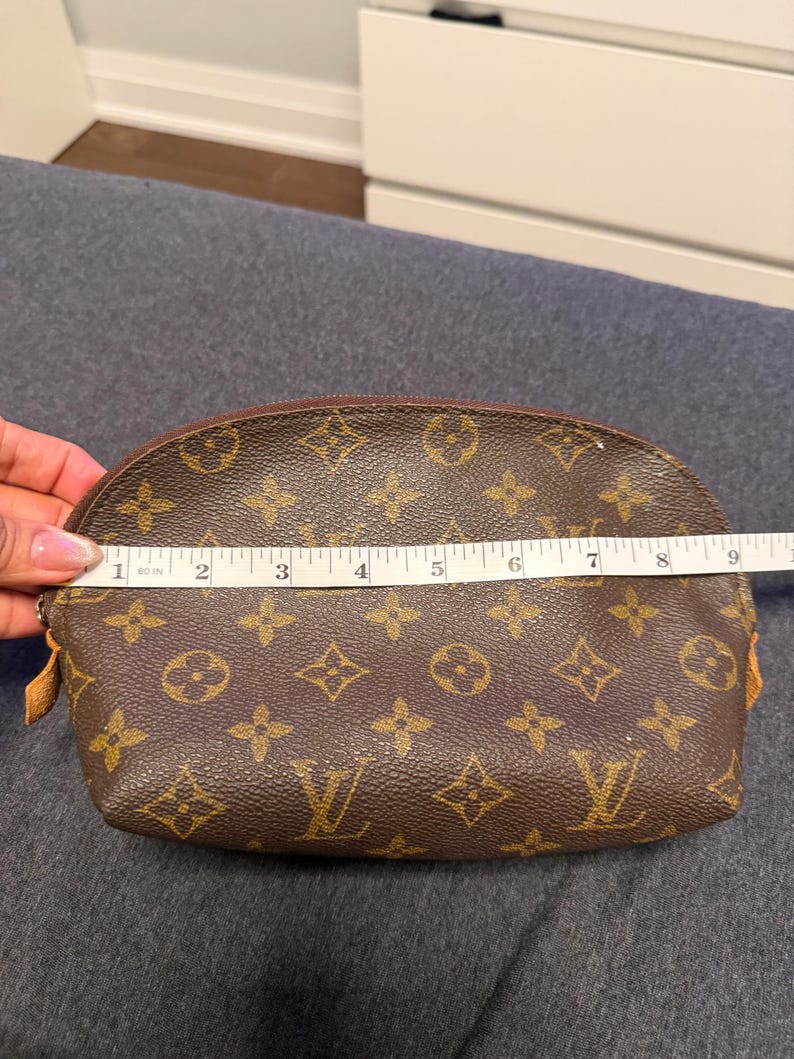 May include: Brown cosmetic bag with a repeating pattern of interlocking floral and logo designs. The bag is approximately 9 inches long. The zipper is brown, and the bag is on a gray surface.