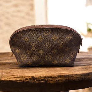 May include: Brown cosmetic bag with a repeating pattern of gold-colored logos. The bag has a curved shape with a brown zipper closure. The bag is sitting on a wooden surface.