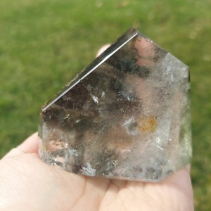 Puede incluir: Un cristal de cuarzo transparente con una inclusión gris ahumado y marrón. El cristal está facetado y tiene forma de pirámide.