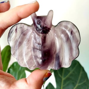May include: A purple and white carved fluorite bat figurine. The bat has a detailed face and wings.