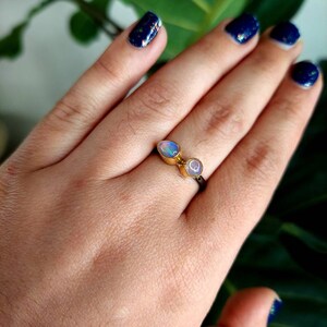 May include: A gold ring with two iridescent opal stones set in a delicate band. The ring is worn on a hand with dark blue nail polish.