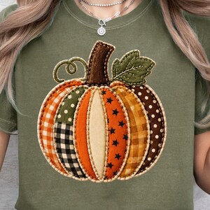 Könnte beinhalten: Ein olivgrünes T-Shirt mit einer großen, mittigen Kürbisgrafik. Der Kürbis besteht aus verschiedenen stoffartigen Flicken in Orange, Braun und Creme und zeigt Muster wie Karos, Sterne und Punkte.