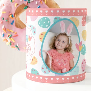 May include: A white ceramic mug with a pink rim and handle. The mug has a colorful Easter egg design with a photo of a child wearing bunny ears in the center. The text "Happy Easter" is printed on the mug.