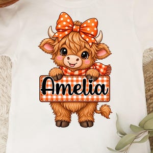 Pode incluir: Uma t-shirt branca de manga curta com o gráfico de uma vaca Highland castanha e felpuda, com um grande laço laranja de bolinhas brancas e um cachecol xadrez a condizer. A vaca segura uma placa retangular com borda laranja e o nome Amelia.