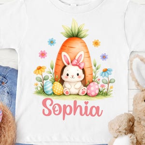 May include: White t-shirt featuring a watercolor illustration of an Easter bunny inside a carrot, surrounded by colorful eggs and flowers. The name "Sophia" is written in pink script. A plush bunny is visible.