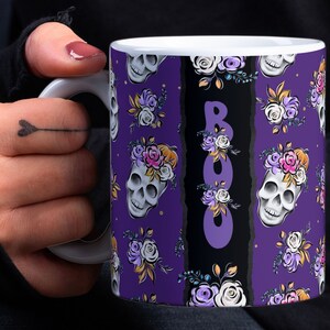 Könnte beinhalten: Eine weiße Keramiktasse mit violettem Hintergrund und einem schwarz-weiß gestreiften Design. Die Tasse zeigt ein sich wiederholendes Muster aus Schädeln mit Blumenkränzen und dem Wort "BOO" in violetten Buchstaben.