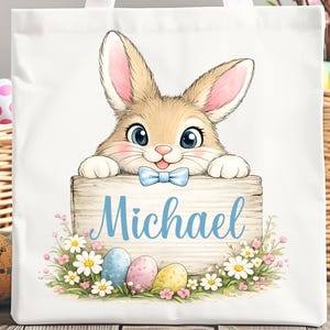 Puede incluir: Bolso de mano blanco con un conejito de dibujos animados sosteniendo un letrero de madera con el nombre "Michael" en escritura azul. El letrero está decorado con flores y huevos de Pascua. Las palabras "Digital Download PNG File" están en la parte inferior.