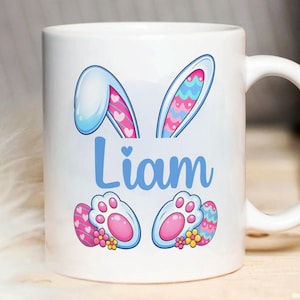 May include: White ceramic mug with an Easter design. It features blue bunny ears, the name "Liam" in blue script, and paw prints with Easter eggs. The ears have pink hearts and blue, pink, and teal patterns.