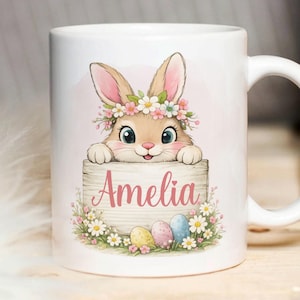 Peut inclure: Mug en céramique blanche avec un lapin de dessin animé portant une couronne de fleurs, tenant un panneau en bois avec le nom "Amelia" en rose. Le design comprend des œufs de Pâques et des fleurs.
