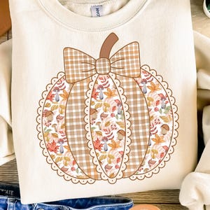 以下が含まれることがあります： 大きなカボチャのデザインが施されたクリーム色のスウェットシャツ。カボチャにはチェック柄のリボンがあり、秋の葉やキノコの模様が詰まっています。画像の下部には「PNG - INSTANT DOWNLOAD」という文字があります。