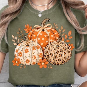 May include: An olive green t-shirt featuring a graphic print of three pumpkins. The central pumpkin has a white heart pattern, the left one has orange flowers, and the right one has a leopard print. Each pumpkin is tied with a tan ribbon bow.