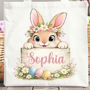 以下が含まれることがあります： 白いトートバッグ。花冠をつけたウサギの水彩イラストで、「Sophia」という名前がピンク色の筆記体で書かれた木の看板を持っています。デザインにはイースターエッグと花が含まれています。「Digital Download PNG File」というテキストが下にあります。