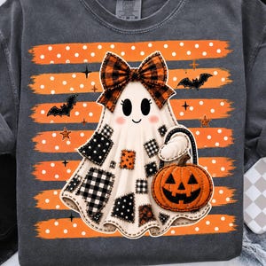 May include: A charcoal grey t-shirt featuring a graphic of a white ghost wearing a plaid bow. The ghost has black and white patterned patches and holds an orange jack-o'-lantern. The background has orange horizontal stripes with white dots.