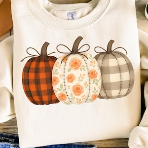 May include: Cream-colored sweatshirt featuring three decorative pumpkins. The pumpkins have different patterns: a plaid design in orange and brown, a floral pattern with orange roses, and a gray and white plaid design. The text "PNG - INSTANT DOWNLOAD" is at the bottom.