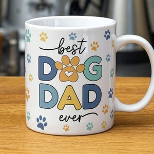 May include: White ceramic mug with the words "best DOG DAD ever" in a playful font. The word "DOG" is in blue and yellow, with a paw print design. Scattered paw prints in blue, yellow, and teal.