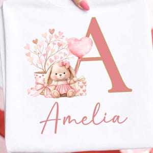 May include: White sweatshirt featuring a pink letter "A" and the name "Amelia". The design includes a bunny wearing a pink dress, a heart-shaped balloon, and a tree with heart-shaped leaves. The words "Digital Download PNG File" are at the bottom.