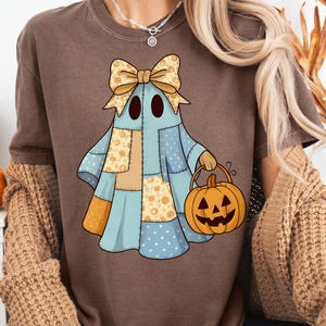 May include: Brown t-shirt featuring a patchwork ghost design with a bow and a jack-o'-lantern bucket. The ghost is in blue, tan, and orange colors. The text "PNG - INSTANT DOWNLOAD" is at the bottom.