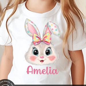 Puede incluir: Una camiseta blanca con un gráfico de conejo de colores y el nombre "Amelia" impreso en rosa. El conejo tiene un lazo rosa con lunares amarillos y una cara blanca con grandes ojos azules.