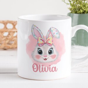 Può includere: Tazza in ceramica bianca con uno sfondo acquerellato rosa che presenta un coniglio dei cartoni animati con un fiocco giallo. Il coniglio ha la pelliccia grigia e gli occhi blu. Il nome "Olivia" è stampato in rosa sotto il coniglio.