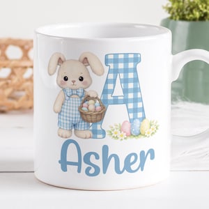 Può includere: Tazza in ceramica bianca con un coniglietto dei cartoni animati che tiene un cestino di uova di Pasqua. La lettera "A" in blu a quadretti e il nome "Asher" sono anch'essi sulla tazza. La tazza ha un manico bianco ed è decorata con grafiche a tema pasquale.