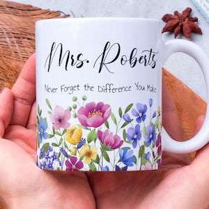 Può includere: Tazza in ceramica bianca con un design floreale e la scritta "Mrs. Roberts - Never Forget the Difference You Make." La tazza presenta un bordo colorato di fiori rosa, viola, gialli e blu.