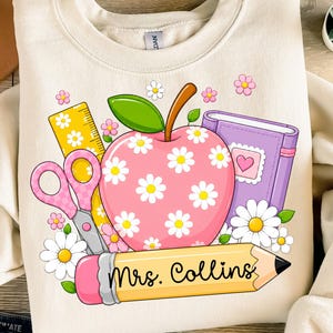 May include: A cream-colored crewneck sweatshirt featuring a graphic print of a pink apple with white daisies, a yellow ruler, pink scissors, a purple notebook with a heart, and a yellow pencil with the text Mrs. Collins printed on it.