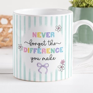 Può includere: Tazza in ceramica bianca con un motivo a righe verticali in azzurro e bianco. La tazza presenta il testo "NEVER forget the DIFFERENCE you make" in lettere colorate, insieme a illustrazioni floreali e un fiocco.