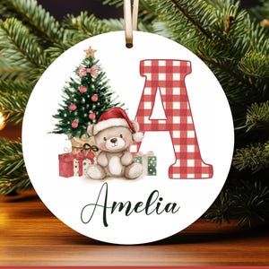 Peut inclure: Ornement de Noël rond avec un motif aquarelle. L'ornement présente un ours en peluche portant un bonnet de Père Noël, un sapin de Noël et la lettre "A" dans un motif à carreaux rouges et blancs. Le nom "Amelia" est écrit en dessous.
