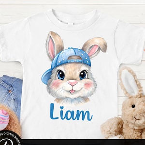 Puede incluir: Una camiseta blanca con un gráfico de un conejo con una gorra de béisbol azul y blanca. El conejo tiene pelaje marrón, ojos azules y una nariz rosa. El texto "Liam" está impreso en azul debajo del conejo.