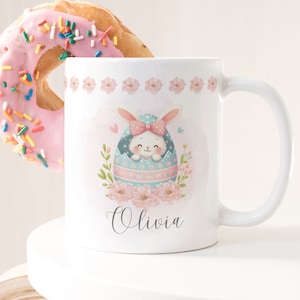 May include: White ceramic mug featuring an Easter bunny design inside a decorated egg, with the name "Olivia" in script. A pink frosted donut with sprinkles sits next to the mug. The mug has a white handle and a floral border.