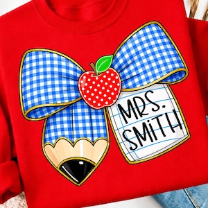 May include: Red sweatshirt featuring a school-themed graphic. The design includes a blue and white gingham bow, a red apple with white polka dots, and a pencil. The text "MRS. SMITH" is written on a notepad.