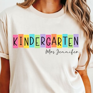 Könnte beinhalten: Cremefarbenes T-Shirt mit dem Wort "KINDERGARTEN" in schwarzen Buchstaben, gesetzt vor einer Reihe farbiger vertikaler Rechtecke. Darunter steht der Name "Mrs Jennifer" in einer handschriftlichen Schriftart.