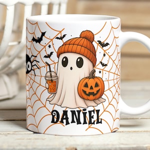 Puede incluir: Taza de cerámica blanca con un diseño de Halloween. El diseño incluye un fantasma de dibujos animados con un gorro naranja y sosteniendo una bebida, una calabaza de Halloween, murciélagos y una telaraña. El nombre "DANIEL" está impreso debajo.