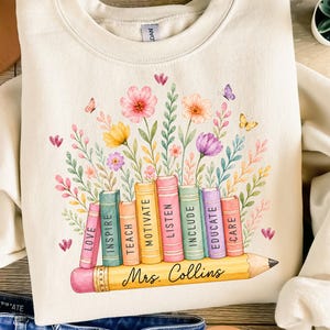 Può includere: Una felpa girocollo color crema con la grafica di una grande matita gialla che sostiene una fila di libri colorati con le etichette Love, Inspire, Teach, Motivate, Listen, Include, Educate e Care, sormontati da fiori e farfalle acquerellate.