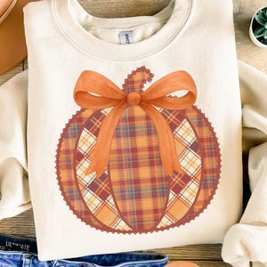 May include: Cream-colored sweatshirt featuring a large pumpkin design. The pumpkin is a plaid pattern in shades of orange, red, and cream, topped with an orange bow. The text "PNG - INSTANT DOWNLOAD" is at the bottom.