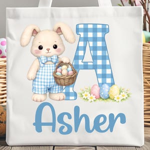 Può includere: Borsa tote bianca con un coniglietto dei cartoni animati in tuta a quadri blu che tiene un cestino di uova di Pasqua. Sono presenti anche una grande lettera "A" a quadri blu e il nome "Asher". Design a tema pasquale.