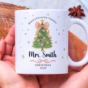 Puede incluir: Taza de cerámica blanca con un diseño de árbol de Navidad, lazo rosa y el texto "Santa's Favorite Teacher" y "Mrs. Smith CHRISTMAS 2025". La taza tiene un asa blanca y estrellas doradas.