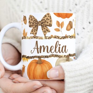May include: White ceramic mug featuring a fall theme with pumpkins, leaves, and acorns. The mug has a bow with a leopard print design and the name "Amelia" in brown script. The mug is held by a person wearing a white sweater.