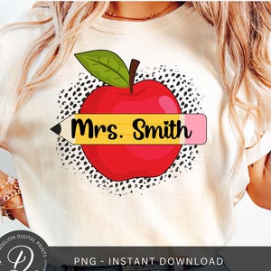 Può includere: Una maglietta bianca con una mela rossa e una matita gialla con il testo "Mrs. Smith" su di essa. La mela è circondata da pois neri e bianchi.