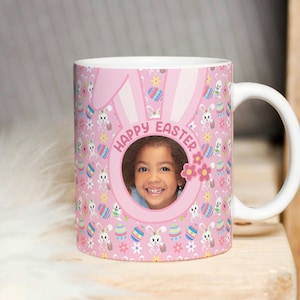 May include: A pink ceramic mug with a bunny ear design and a photo frame in the centre. The mug has a white handle and is decorated with a pattern of Easter eggs, bunnies, and flowers. The text "Happy Easter" is written around the photo frame.