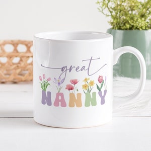 Puede incluir: Taza de cerámica blanca con un diseño floral y el texto "great nanny" en escritura morada. Las letras de "nanny" están en colores pastel y cada letra tiene una flor diferente que crece fuera de ella.